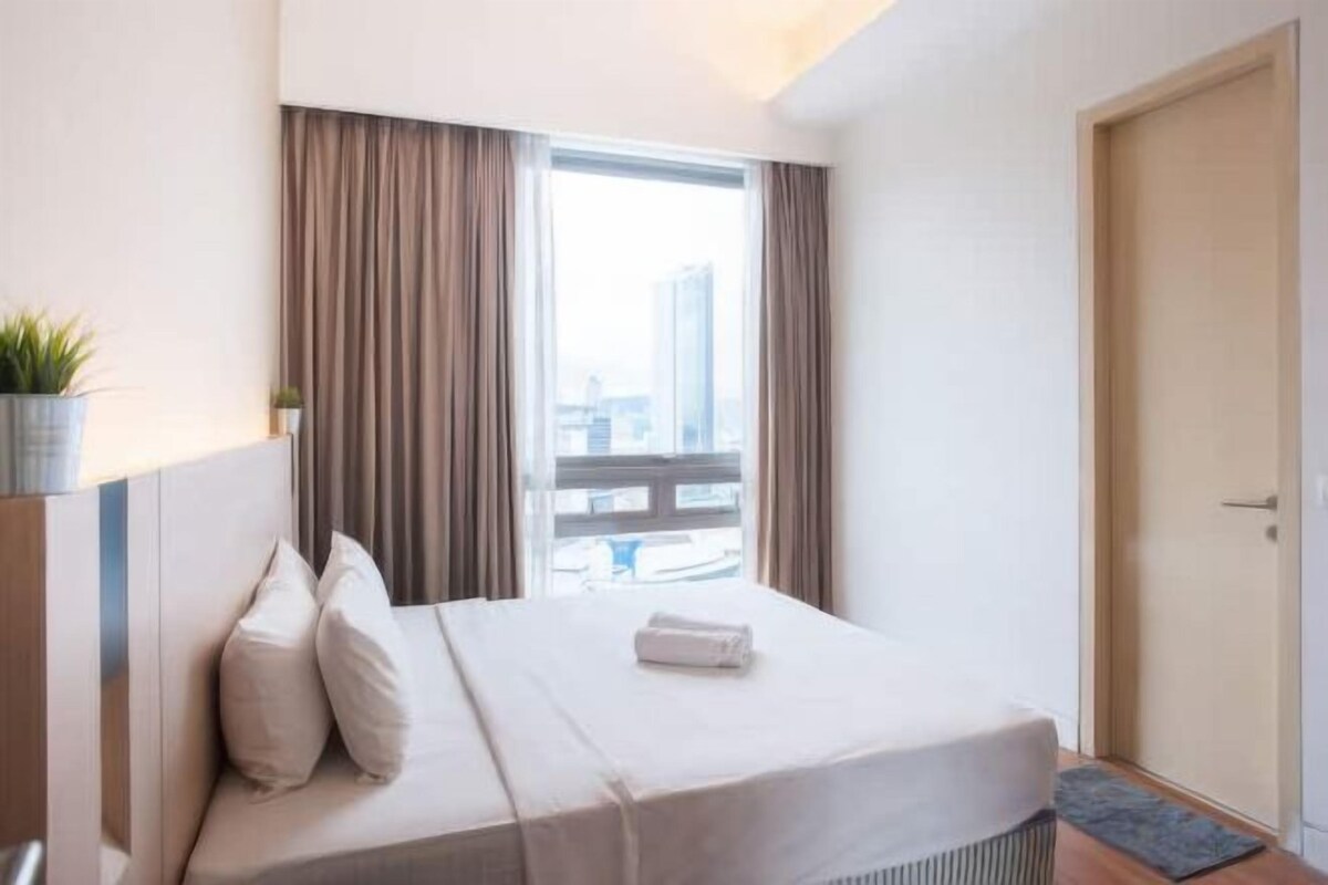 A hotel room with a bed, a window with curtains, a door, and a view of buildings outside.