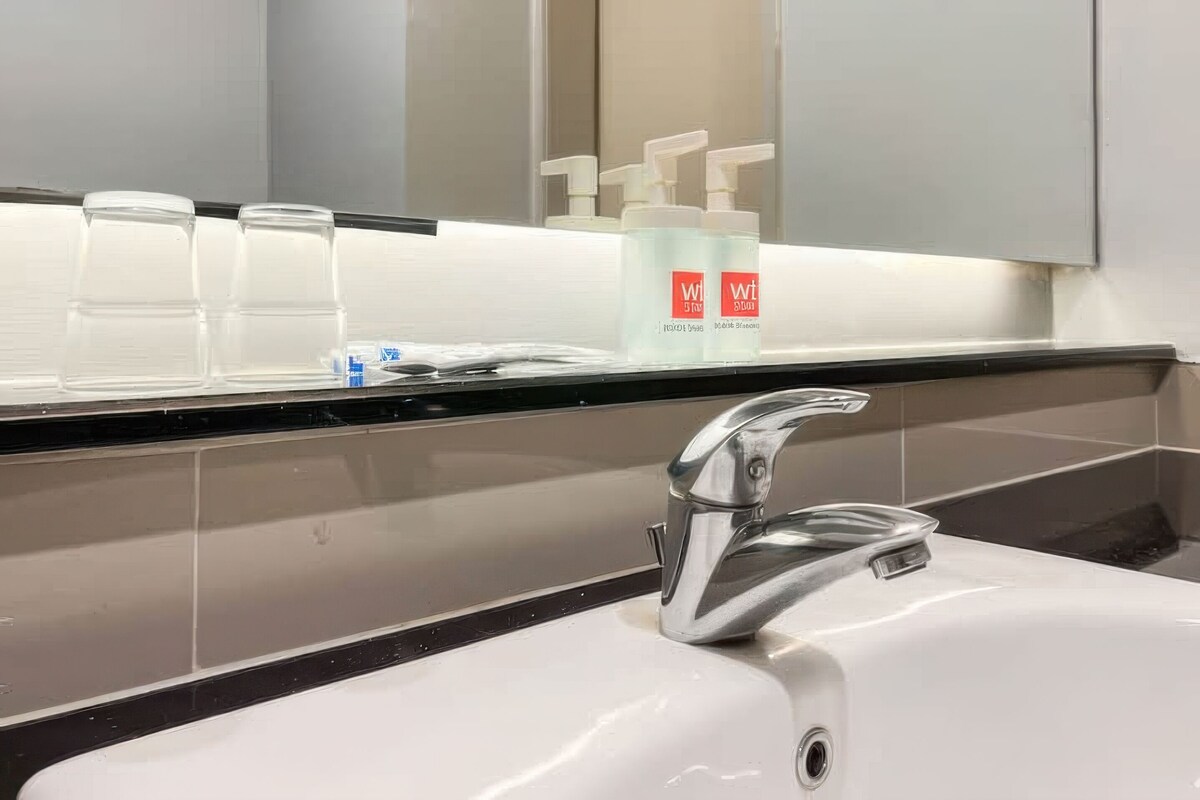 Une salle de bain moderne avec un lavabo blanc, un robinet chromé et un miroir au-dessus du lavabo. On y trouve deux gobelets en plastique transparent et deux flacons blancs dotés de pompes doseuses, posés sur le comptoir.