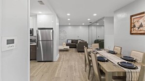 Una cocina moderna con refrigerador y microondas de acero inoxidable, una zona de comedor con mesa de madera para cuatro personas y una sala de estar con sofá y un elemento decorativo en la pared.