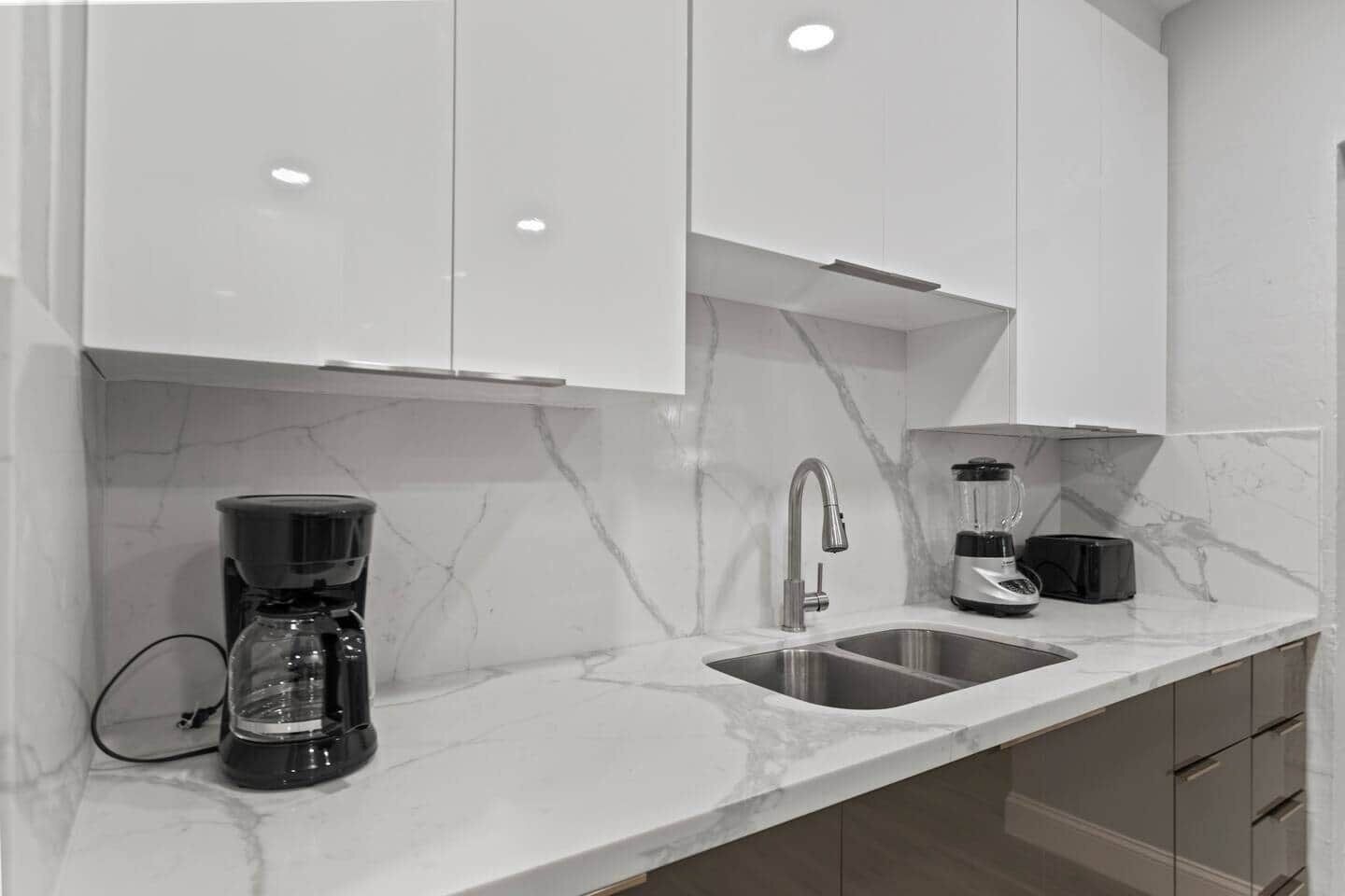 A modern kitchen with white cabinets, a marble countertop, a coffee maker, a blender, and a sink.
