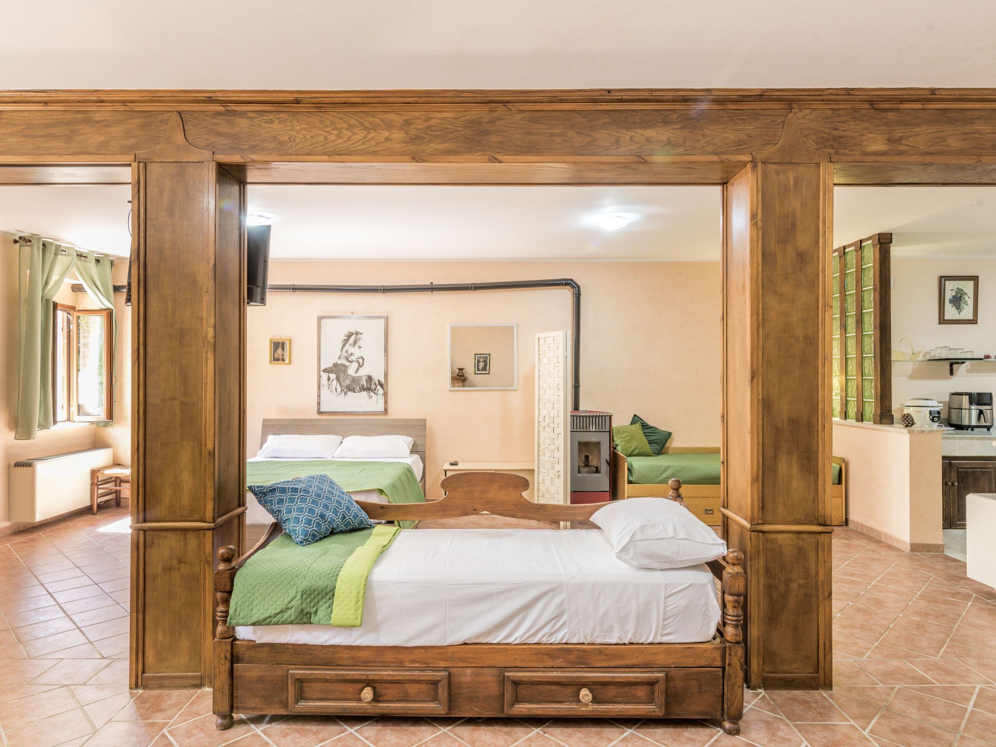 A bedroom with a wooden bed, a small bedside table, a nightstand, a green chair, a window with curtains, a framed picture on the wall, and a kitchen area with a stove and cabinets.