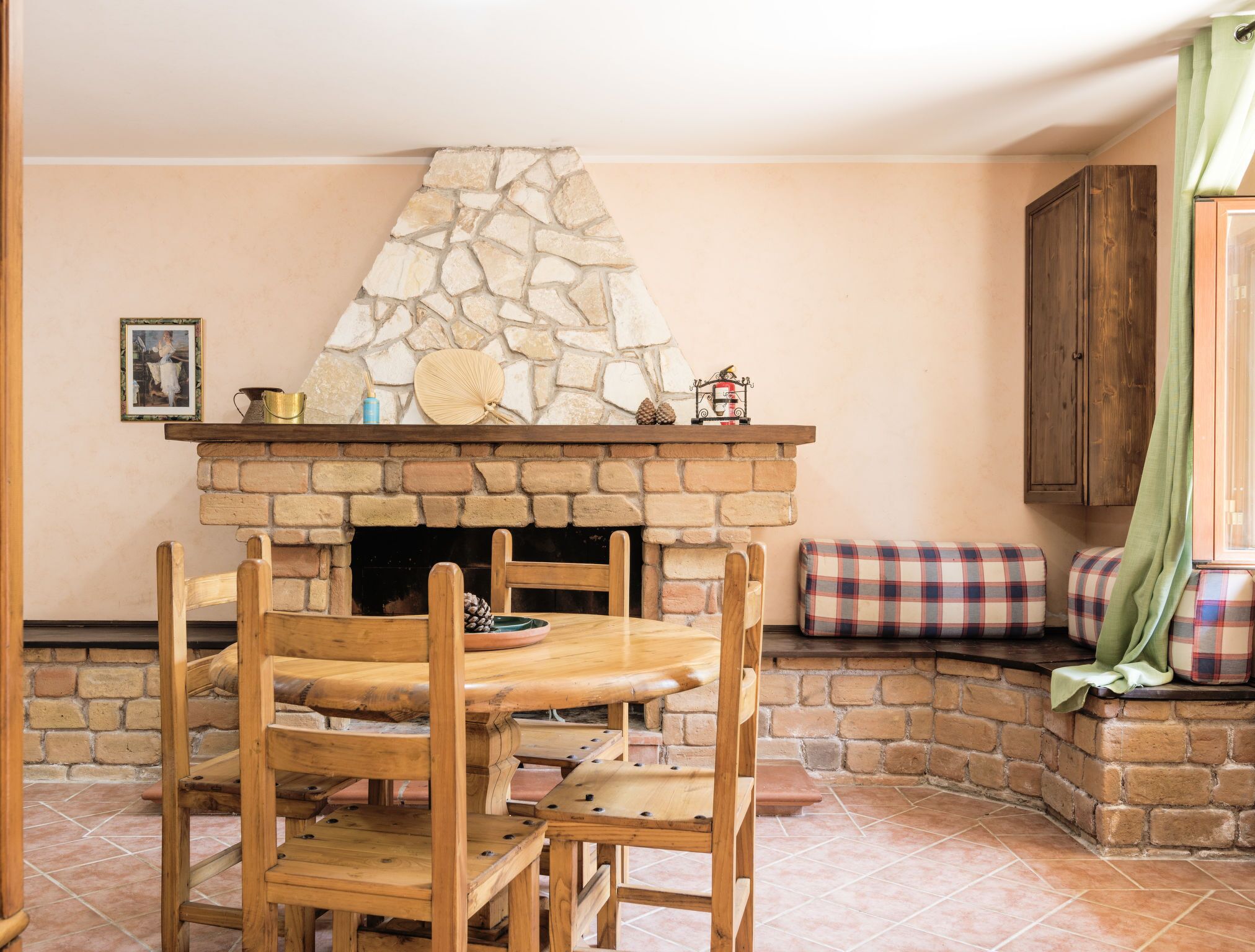 Una chimenea de piedra con repisa de madera, una mesa de comedor de madera con sillas, y un cojín a cuadros en un banco.