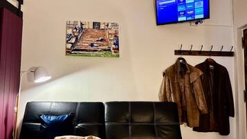 A room with a black leather couch, a mounted TV displaying streaming services, a wall-mounted coat rack with jackets, and a framed picture of a staircase.