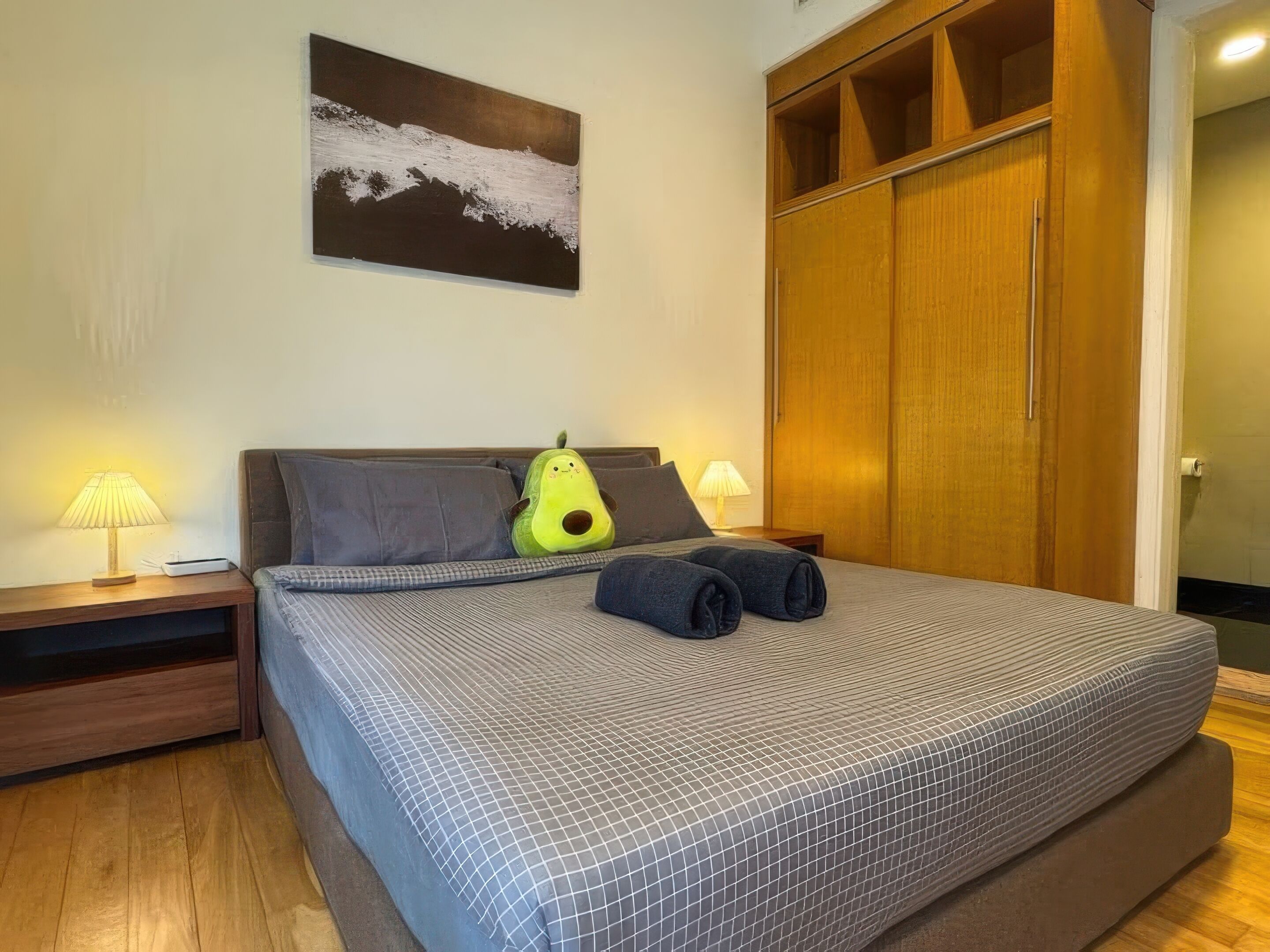 A neatly made bed with a patterned bedspread, two pillows, a green pillow, and rolled towels. There are two bedside lamps, a wooden nightstand, and a wooden wardrobe with a mirror.