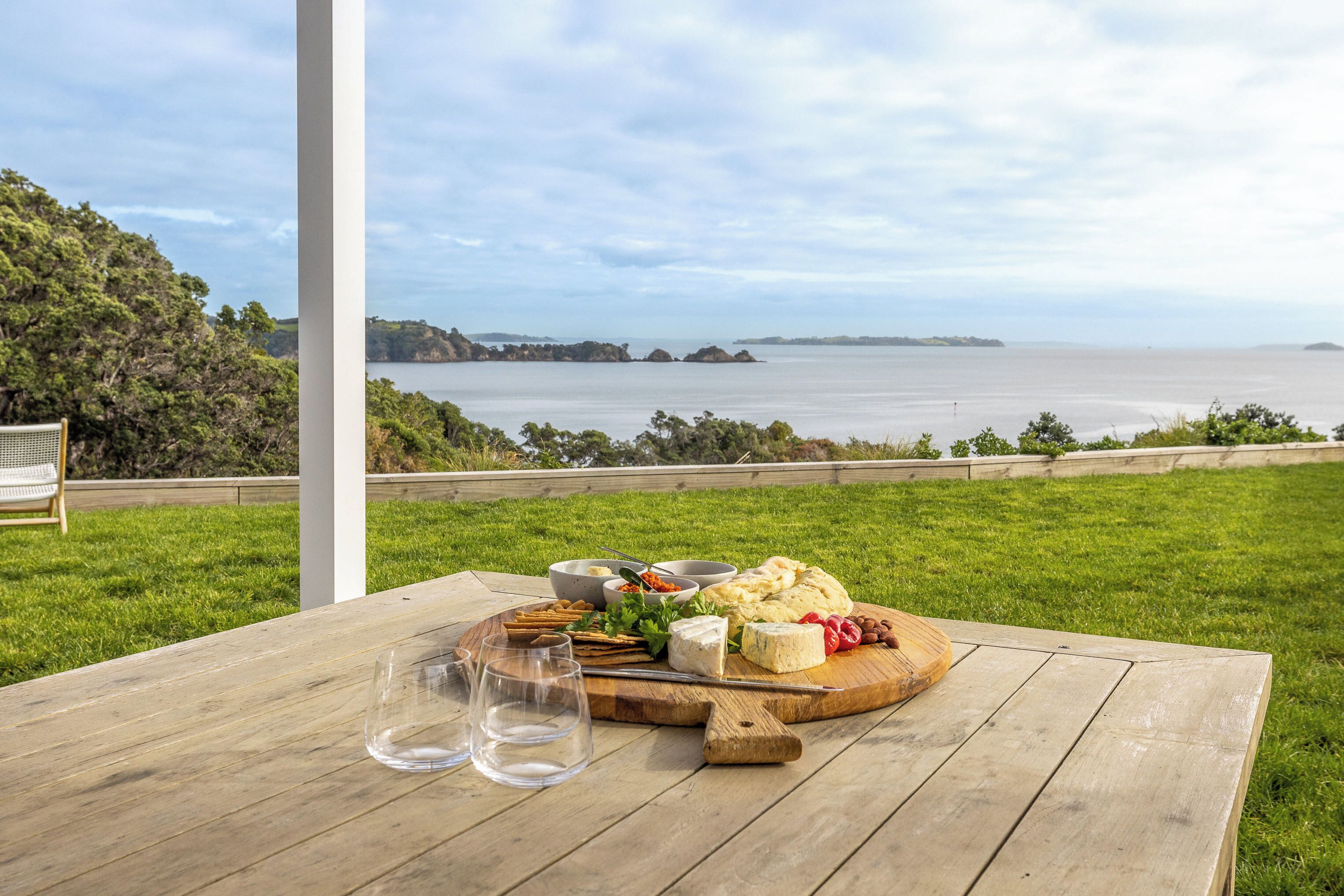 A wooden table with a cheese platter, glasses, and a scenic view of the ocean.