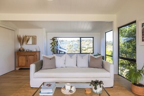 A modern living room with a white sofa, a coffee table with books and decorative items, a wooden cabinet, and large windows offering a view of a hillside.