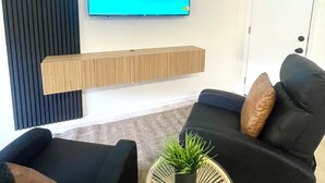 Una sala de estar moderna con un televisor de pantalla montado mostrando el logotipo de Disney+, un estante de madera, dos sofás negros con cojines y una pequeña planta en maceta sobre una mesita redonda.