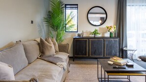 Ein modernes Wohnzimmer mit einem beige farbenen Sofa, einer dunklen Holzkommode, einem runden Spiegel und einem Couchtisch mit Büchern.