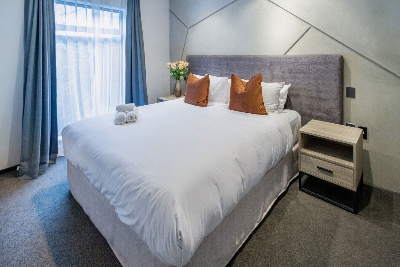 A hotel room with a large bed, a nightstand, a vase with flowers, and a window with curtains.