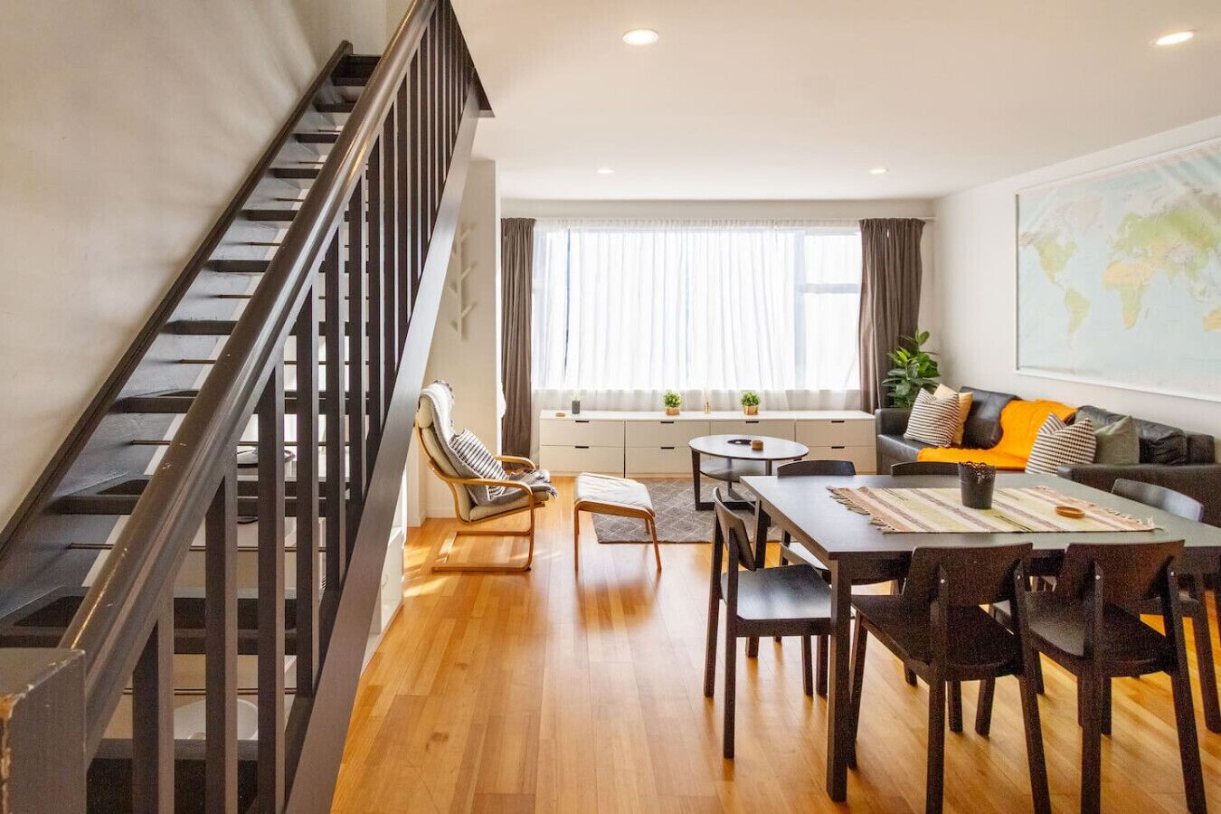 A modern living room with a dining area, a staircase, and a large window with curtains.
