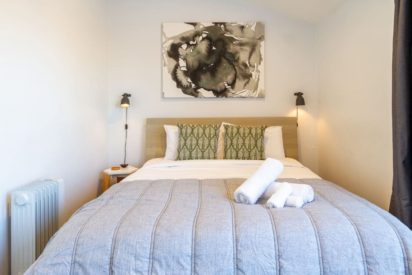 Une chambre d’hôtel avec un lit, un radiateur, une table de chevet, une lampe et un grand tableau abstrait accroché au mur.