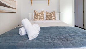 A neatly made bed with a blue quilt, two patterned pillows, and a rolled white towel on the bed.