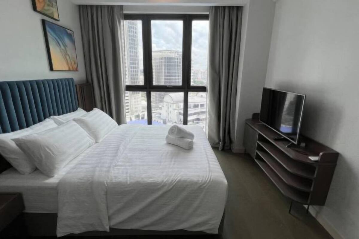 A hotel room with a bed, a television, a window with curtains, and a view of buildings.