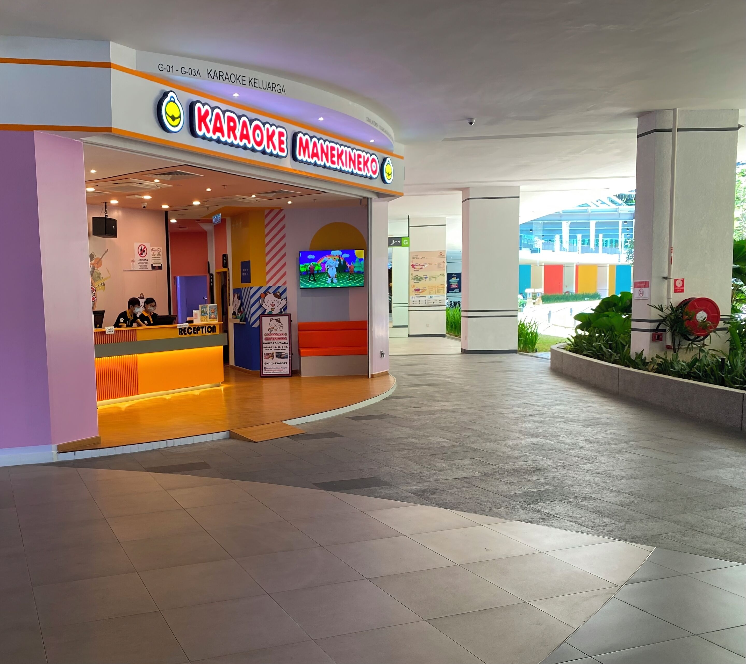 A karaoke area with a reception desk, a TV screen displaying a cartoon, and a sign indicating it's in G-01 - G-03.