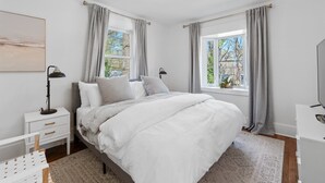 A bedroom with a large bed, two bedside tables, a nightstand, a desk, and a television. - Greenville Bungalow Near Downtown + Swamp Rabbit (Greenville)