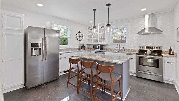 A modern kitchen with a stainless steel refrigerator, double ovens, a built-in island with bar stools, white cabinetry, and pendant lighting. - Greenville Bungalow Near Downtown + Swamp Rabbit (Greenville)