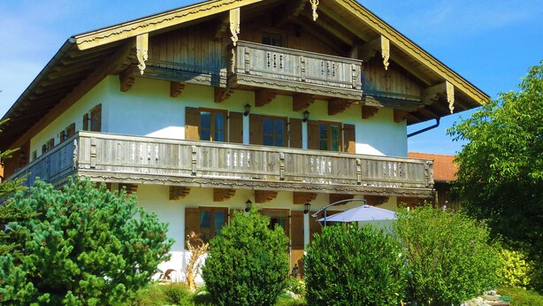 Een traditionele chalets met een houten balkon, een balkon op de tweede verdieping, en een tuin met bomen en een bankje.