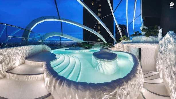 Ein modernes Spa mit einer zentralen Whirlwanne, umgeben von Sitzbereichen, mit Blick auf eine Stadtlandschaft und einem markanten, blau beleuchteten Gebäude.