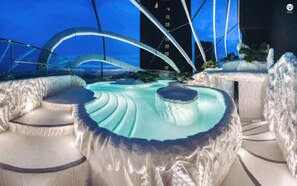 Un spa moderne doté d’un grand bain à remous central, entouré d’espaces de détente, le tout mis en valeur par une vue sur la ville et une structure remarquable illuminée de lumière bleue.