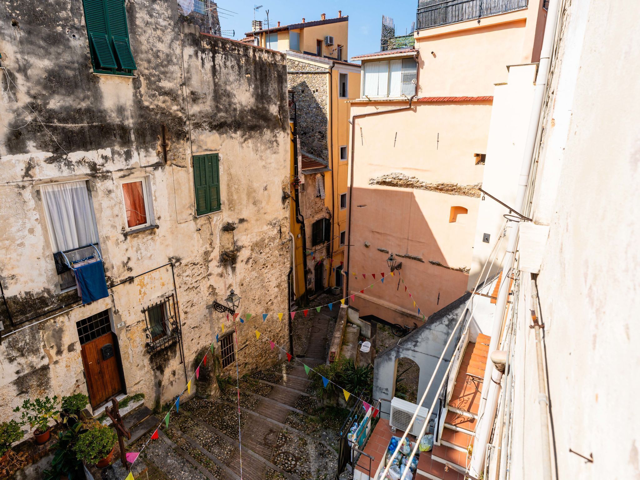 Une ruelle étroite entre deux bâtiments, avec une petite cour, des marches en pierre et quelques plantes en pot.