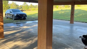 Un espacio al aire libre cubierto con suelo de hormigón, pilares de madera, un ventilador de techo y una parrilla.