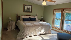 Un dormitorio con cama, ventilador de techo, mesita de noche, lámpara, un cuadro enmarco y ventana con vistas a árboles.
