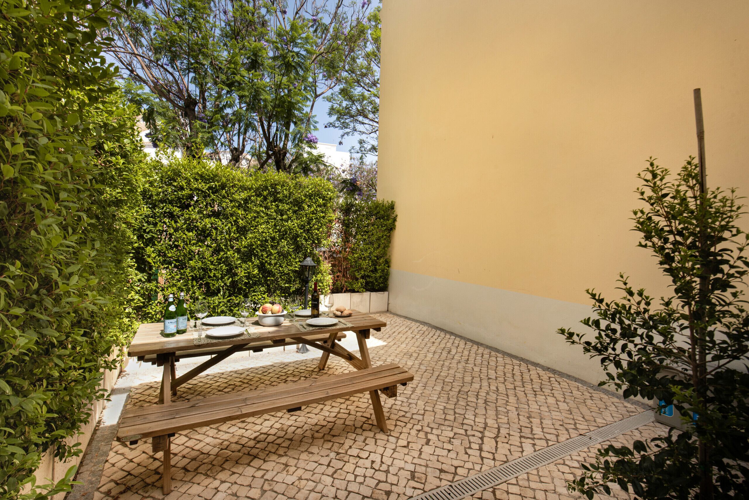 A wooden picnic table with benches, surrounded by greenery and a paved patio area.