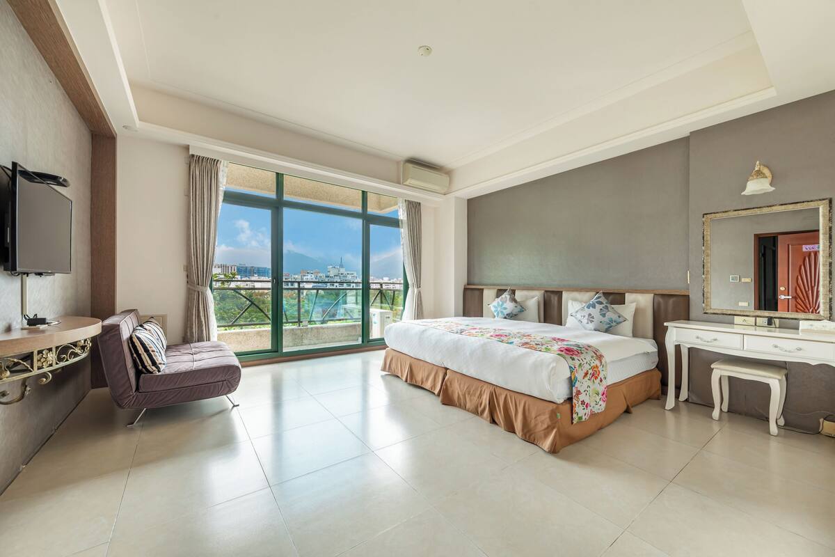 Ein Hotelzimmer mit einem großen Bett, einem Schreibtisch, einem Sessel, einem Fernseher und einem Balkon mit Blick auf die Stadt.