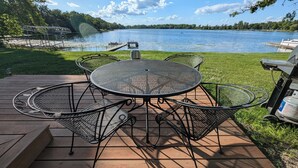 Une terrasse dotée d’une table et de chaises en métal, de forme ronde, qui offre une vue sur un lac avec un quai et un bateau.