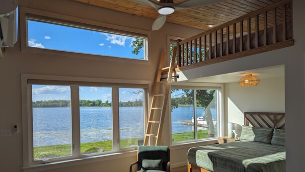 Une chambre spacieuse dotée d’une grande fenêtre donnant sur un lac, d’un plafond en bois et d’une mezzanine avec vue sur l’eau.