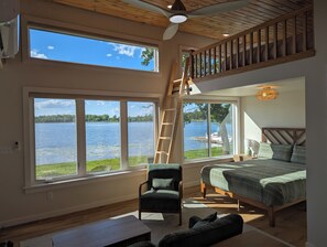 Une chambre spacieuse dotée d’une grande fenêtre donnant sur un lac, d’un plafond en bois et d’une mezzanine avec vue sur l’eau.