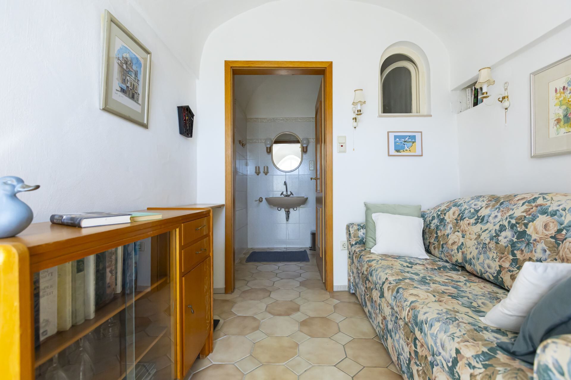 Una habitación acogedora con un escritorio de madera, un sofá floral y un baño con lavamanos y espejo.