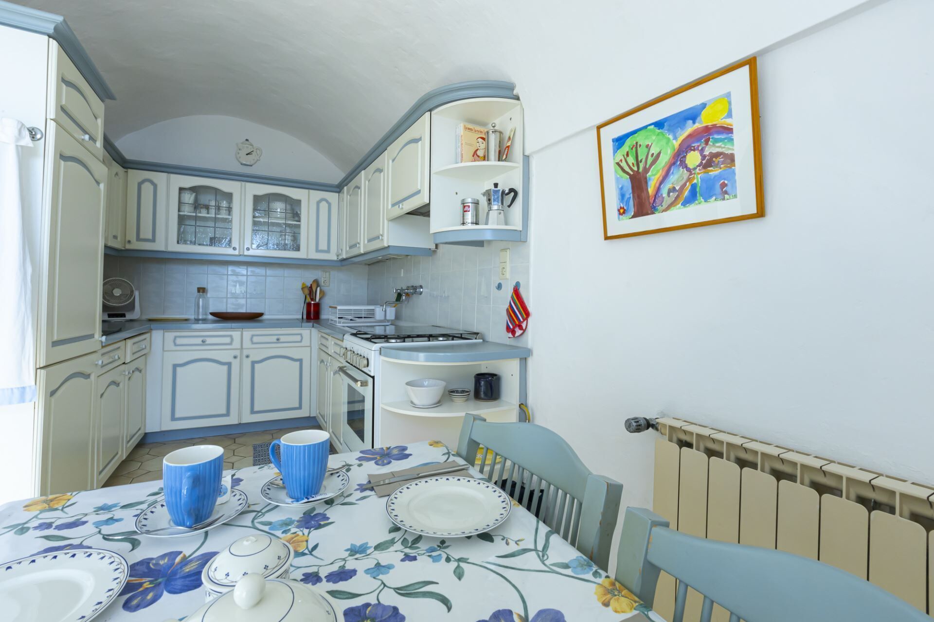 Een keuken met witte en blauwe kastjes, een fornuis en een tafel gedekt met blauwe bekers en borden.