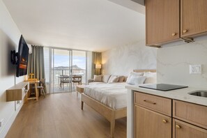 A modern hotel room with a large bed, a desk with a chair, a television, and a balcony with a view of the city.