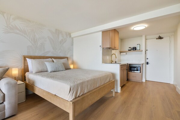 A bedroom with a wooden bed, a bedside table, a chair, a small desk with a chair, a kitchenette, and a door.