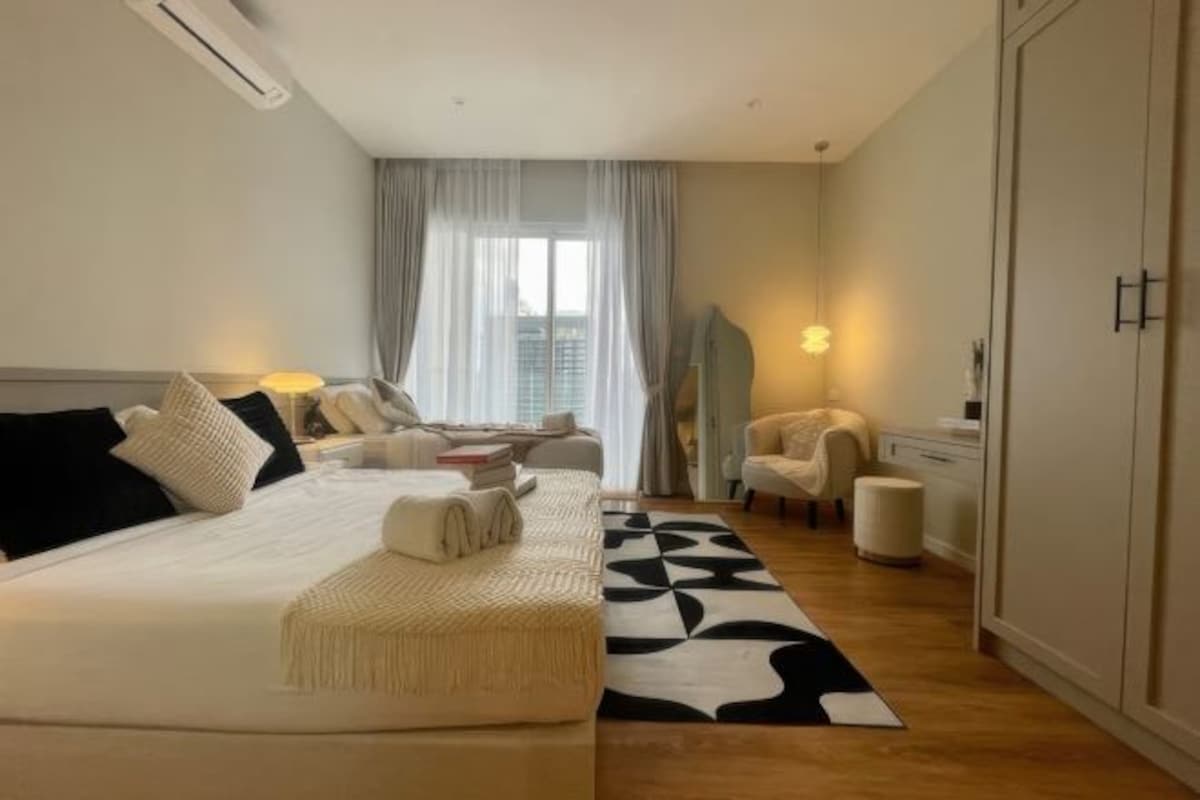 Une chambre d’hôtel moderne avec un grand lit, un canapé, un fauteuil et un bureau. On y trouve une fenêtre avec des rideaux, une lampe et un climatiseur fixé au mur.