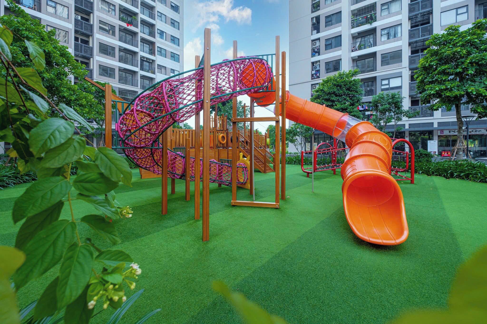 A playground with a slide and climbing structures, surrounded by greenery and residential buildings.