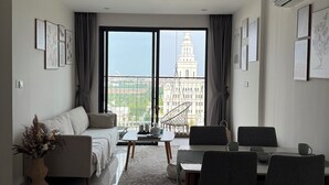A modern living room with a sofa, dining table, and chairs, large windows with a view of a building, and artwork on the walls. - Moon Homestay 96 (Hanoi)