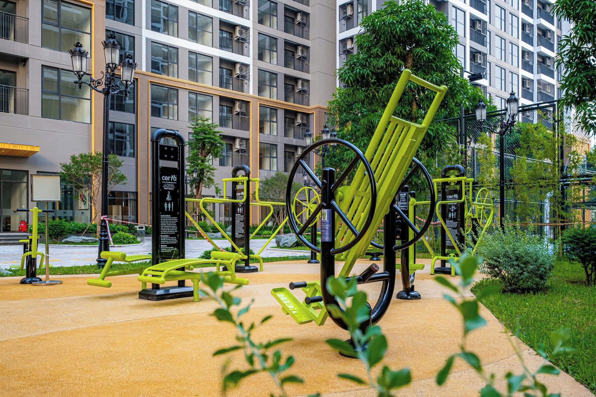 An outdoor gym with exercise equipment, surrounded by greenery and a modern building in the background.