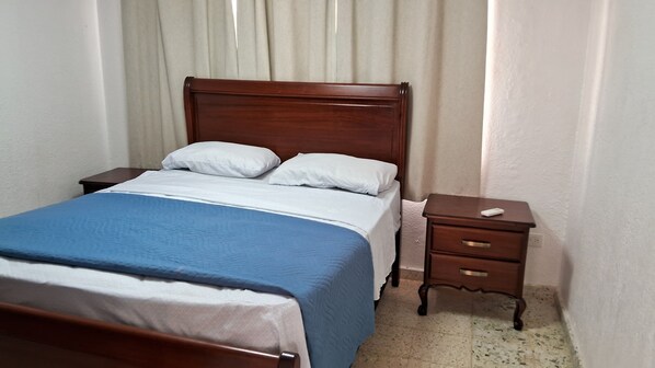 A neatly made bed with a blue bedspread and white pillows, a wooden nightstand with a drawer, and a window with white curtains.