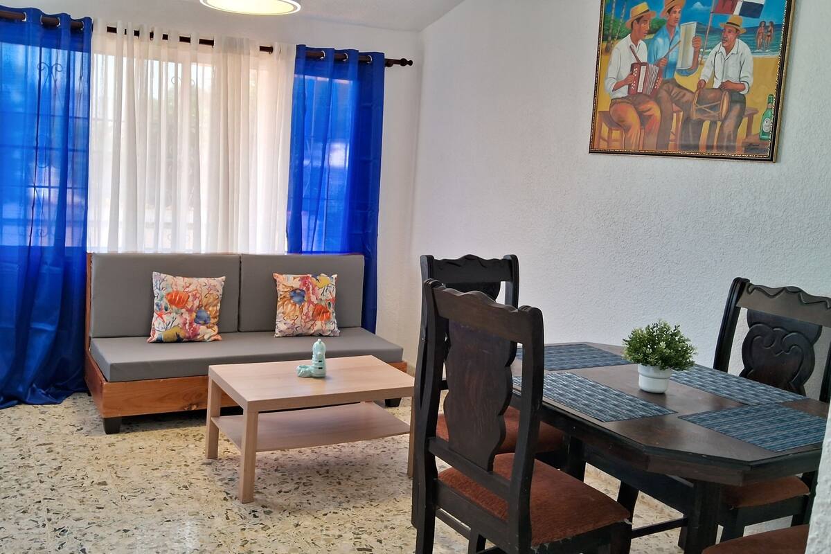 A cozy living area with a sofa, two colorful pillows, a small table, and a dining table with chairs.