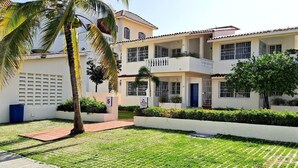 A two-story building with a white facade, multiple balconies, and a paved courtyard with grass and palm trees. - CortLang Vacation Rentals Costambar (Puerto Plata)
