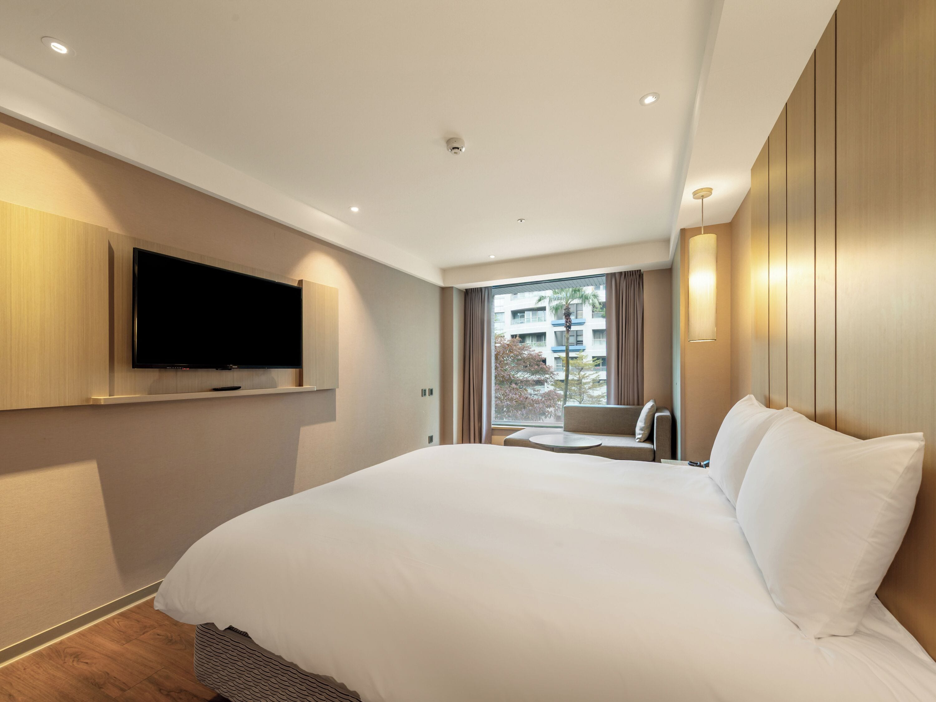 Una habitación de hotel moderna con una cama grande, un televisor de pantalla plana en la pared, un pequeño rincón de descanso con una mesa y un amplio ventanal con vistas a la ciudad.