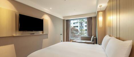 Una habitación de hotel moderna con una cama grande, un televisor de pantalla plana en la pared, un pequeño rincón de descanso con una mesa y un amplio ventanal con vistas a la ciudad.