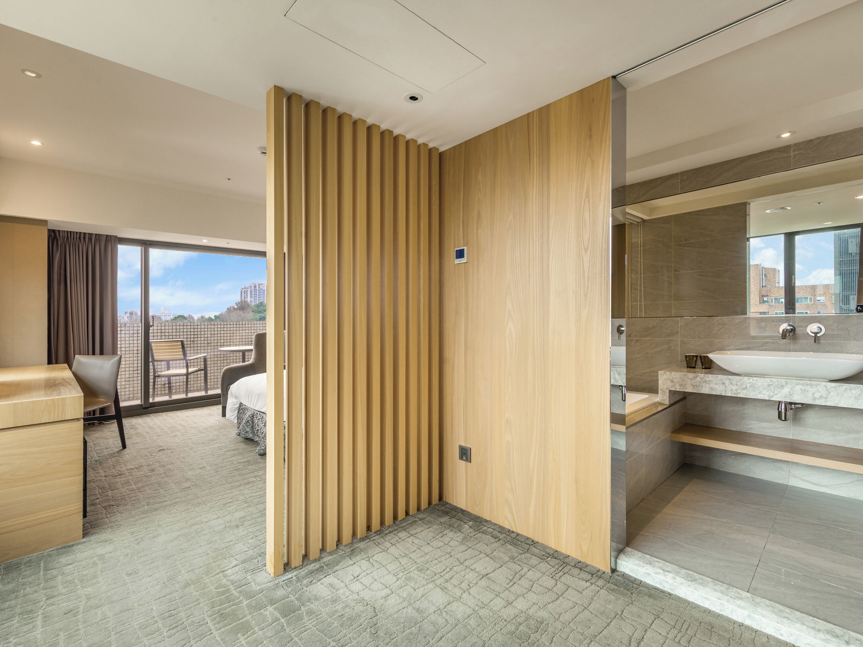 A modern hotel room with a balcony, a wooden partition, a bathroom with a large sink and mirror, and a view of buildings.