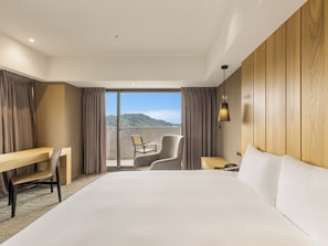 A hotel room with a large bed, a desk, a chair, and a view of a mountainous landscape through a sliding glass door.