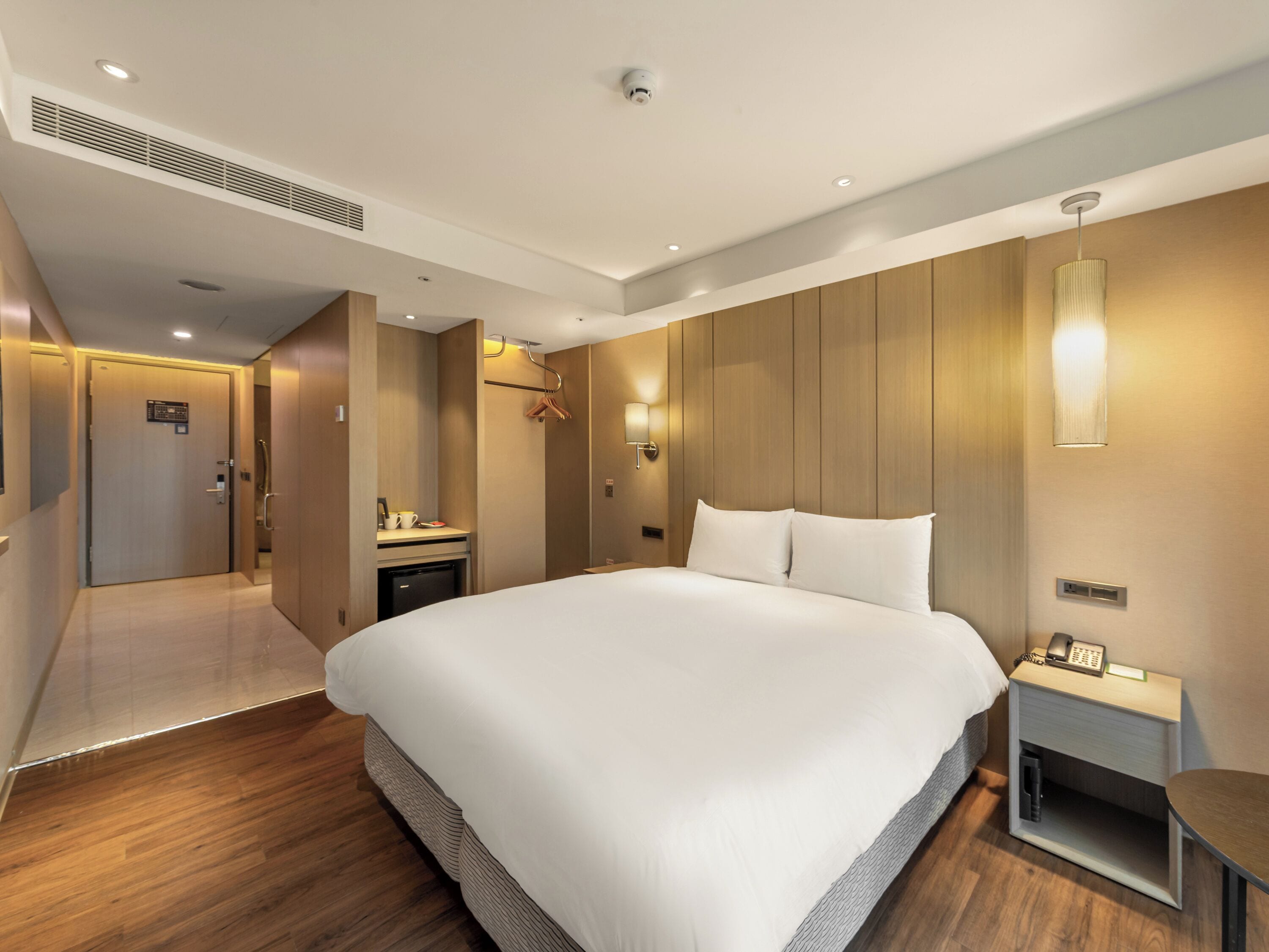 Habitación de hotel moderna con una cama grande, una mesita de noche con un teléfono, una lámpara fijada a la pared y una puerta que da a un baño.