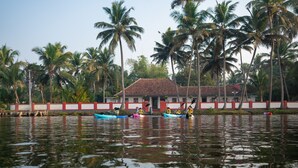 A calm water body with kayaks and a building with a red roof. - Kuttanad Kayak Club- Riverside Heritage Homestay In Alleppey Backwater village (Alappuzha)