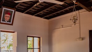 A room with a red floor, two blue cushioned chairs, a woven rug, a ceiling fan, a wooden door, and a window with bars. - Kuttanad Kayak Club- Riverside Heritage Homestay In Alleppey Backwater village (Alappuzha)