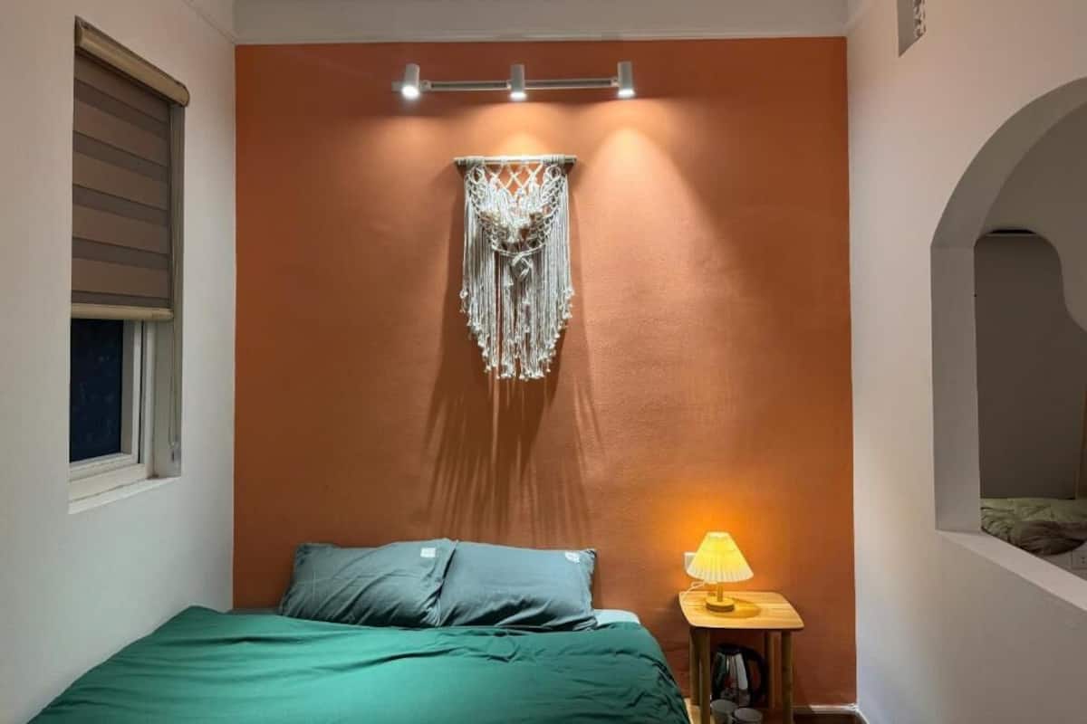 A bedroom with a bed, a wall-mounted macrame piece, a lamp, and a window with blinds.